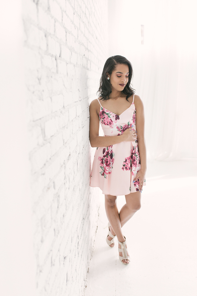 Softly posed image of Connie looking down, standing by a white wall in her pink floral dress and heels at Reflection Images by Tracy Rowell in Florence SC