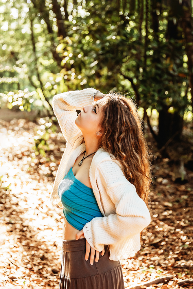 Side profile of Eve in turquoise top and cream sweater, hand in hair, golden forest light…