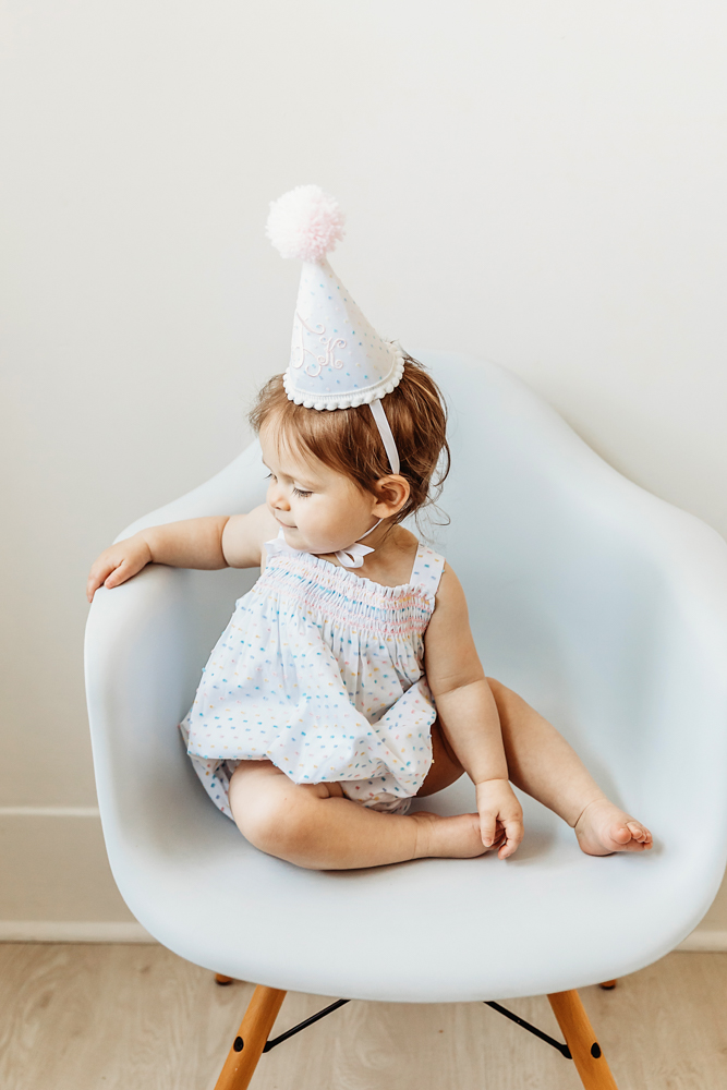 Birthday girl sitting in a modern chair wearing a pastel birthday hat, looking to the side.