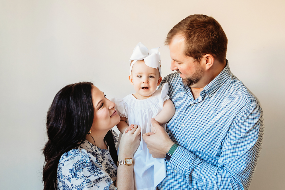 Elsie smiling in her parents’ arms, wearing a white dress and bow, soft studio light at Reflection Images by Tracy Rowell in Florence SC