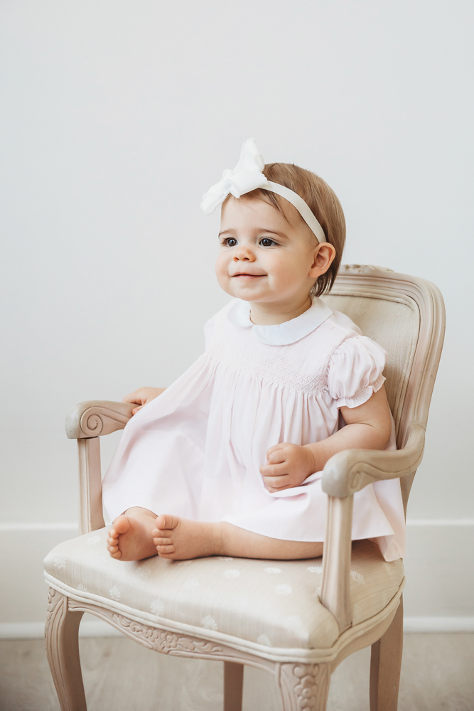 Grace sitting barefoot on a vintage cream chair in a pale pink dress and white bow at Reflection Images by Tracy Rowell in Florence SC