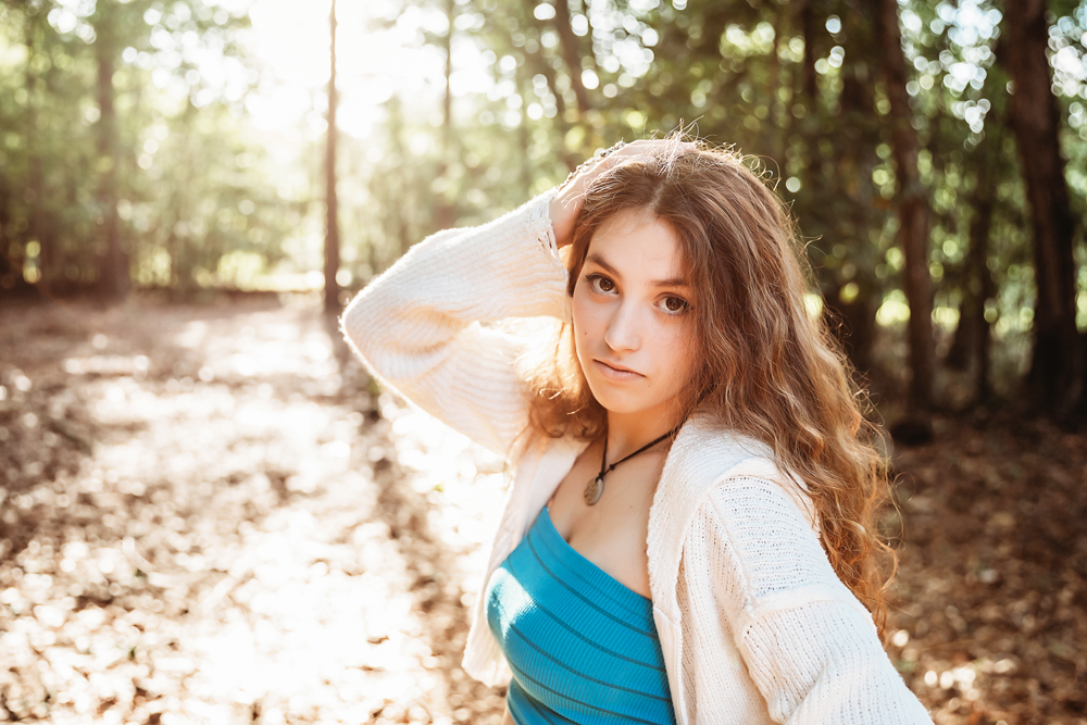 Eve looking into camera with hand in hair, turquoise top, natural forest backdrop…