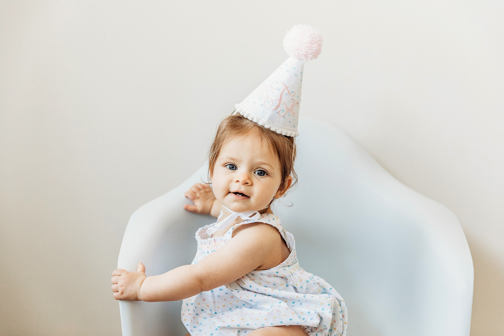 One-year-old girl in birthday hat turning her head toward the camera.