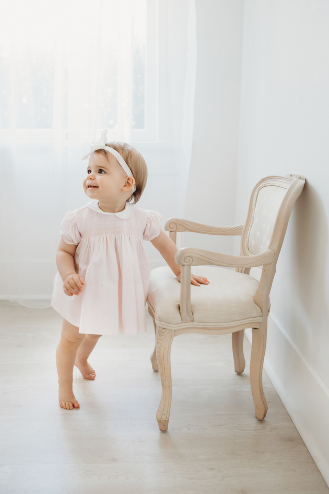 Grace standing beside a neutral-toned chair, softly lit by window light, wearing a pale pink dress and bow at Reflection Images by Tracy Rowell in Florence SC