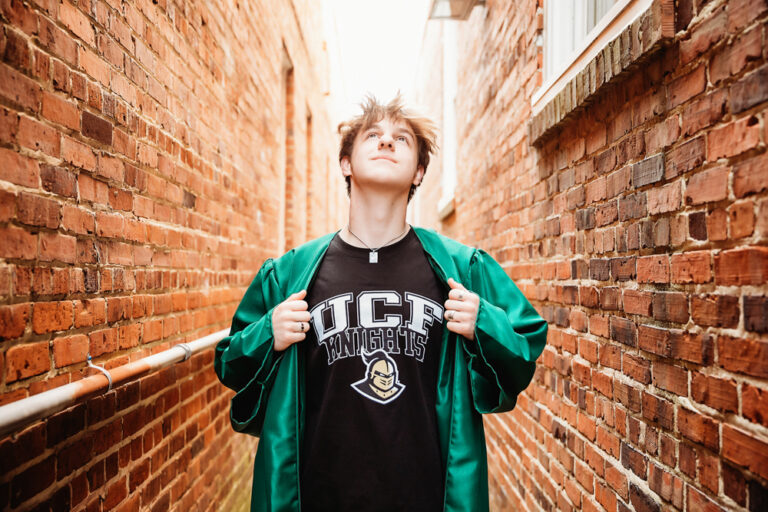 Creative graduation portrait of Jack pulling open his green gown to reveal a UCF Knights shirt underneath, standing in a brick alleyway at Reflection Images by Tracy Rowell