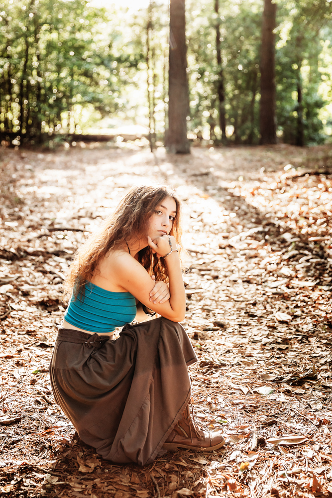 Eve sitting on leafy ground in turquoise top and brown skirt, sunlight filtering through trees…