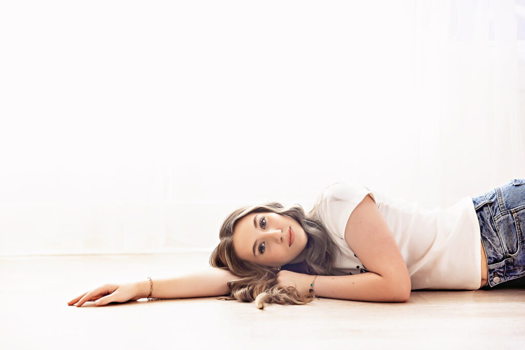 Addy lying on the light wood floor, head resting on her arm, gazing warmly toward camera with sheer curtain light at Reflection Images by Tracy Rowell in Florence SC
