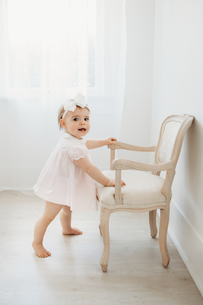 Grace turning back with a smile while standing and holding onto a vintage chair in window light at Reflection Images by Tracy Rowell in Florence SC