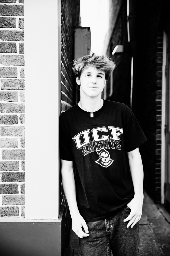 Black and white portrait of Jack leaning against a brick wall in a UCF Knights shirt during his senior session in downtown Florence SC with Reflection Images