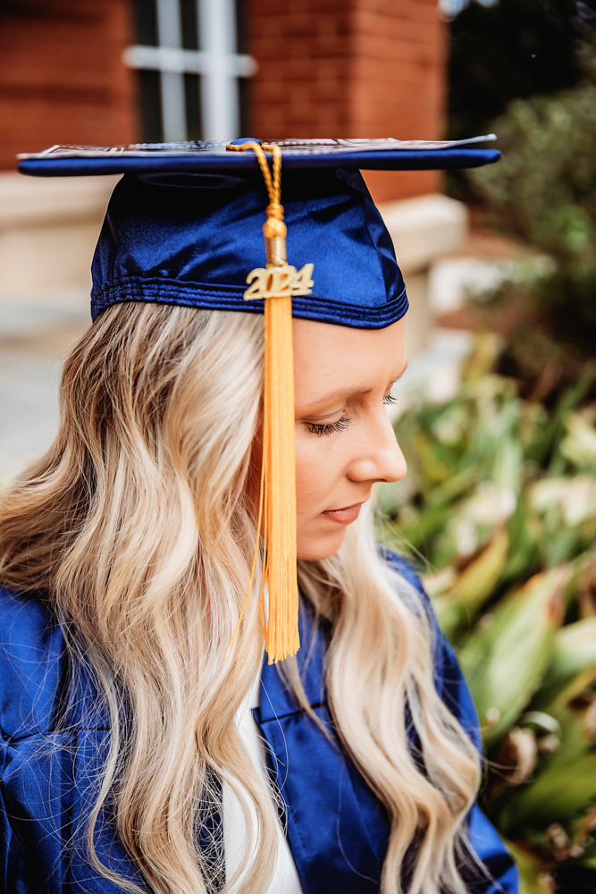 Kristina sitting gracefully on stone steps in her navy graduation gown with a brick wall and greenery in the background at Reflection Images by Tracy Rowell in Florence SC