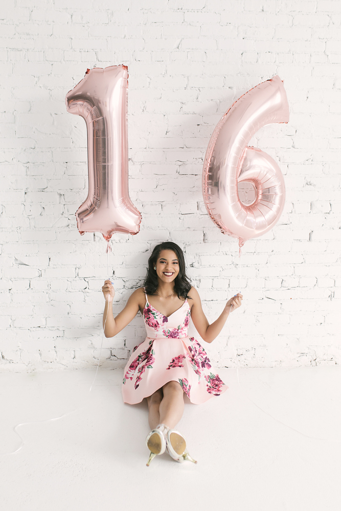 Connie sitting cross-legged on the floor holding rose gold 16 balloons, smiling in her floral dress at Reflection Images by Tracy Rowell in Florence SC