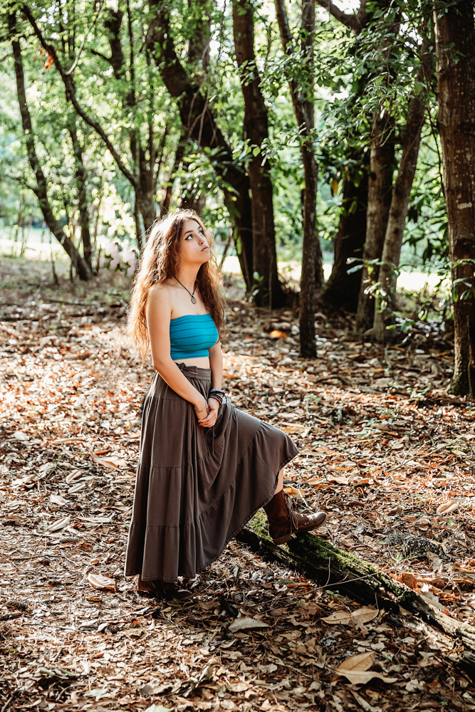 Eve standing with one foot raised on a tree root, turquoise top and brown skirt, gazing upward…