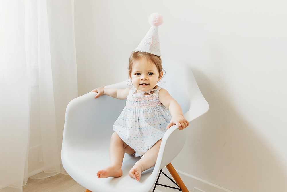 Toddler smiling and sitting confidently in white chair wearing a birthday hat.
