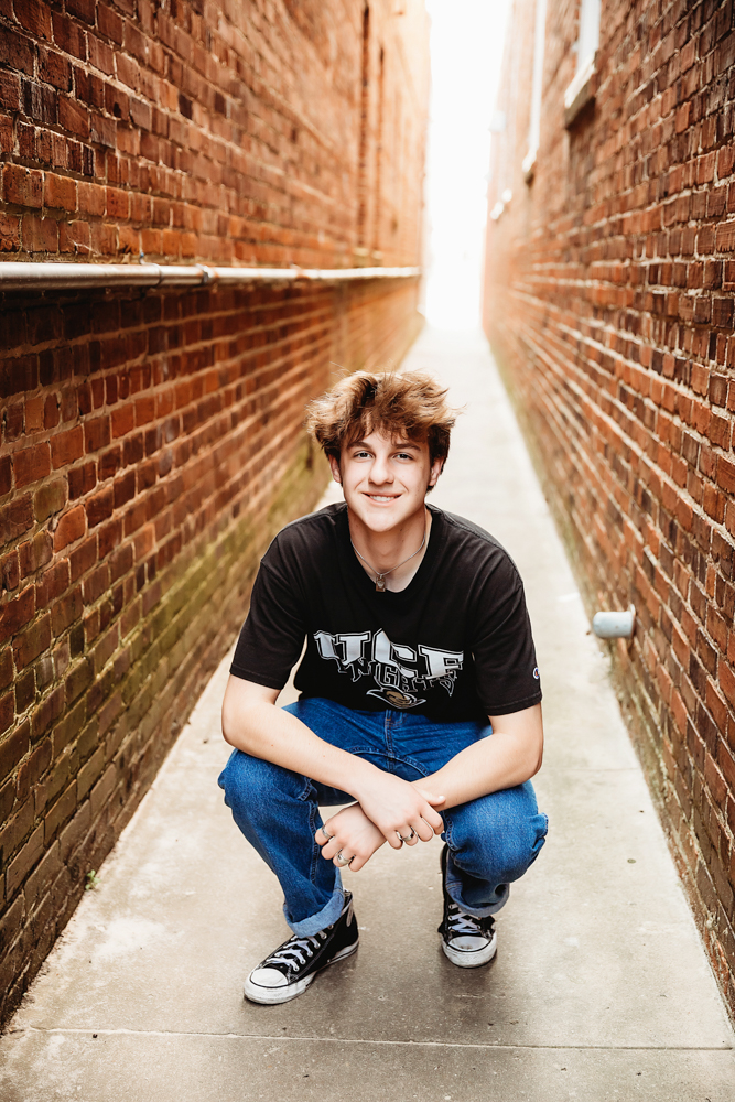 Jack squatting in a relaxed pose with rolled jeans and Converse in a vintage brick alley during his Florence SC senior session with Reflection Images