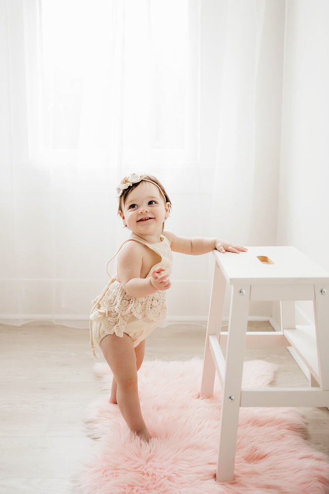 Aubrey standing on a blush pink rug next to a white stool, in a cream lace romper and flower headband at Reflection Images by Tracy Rowell in Florence SC