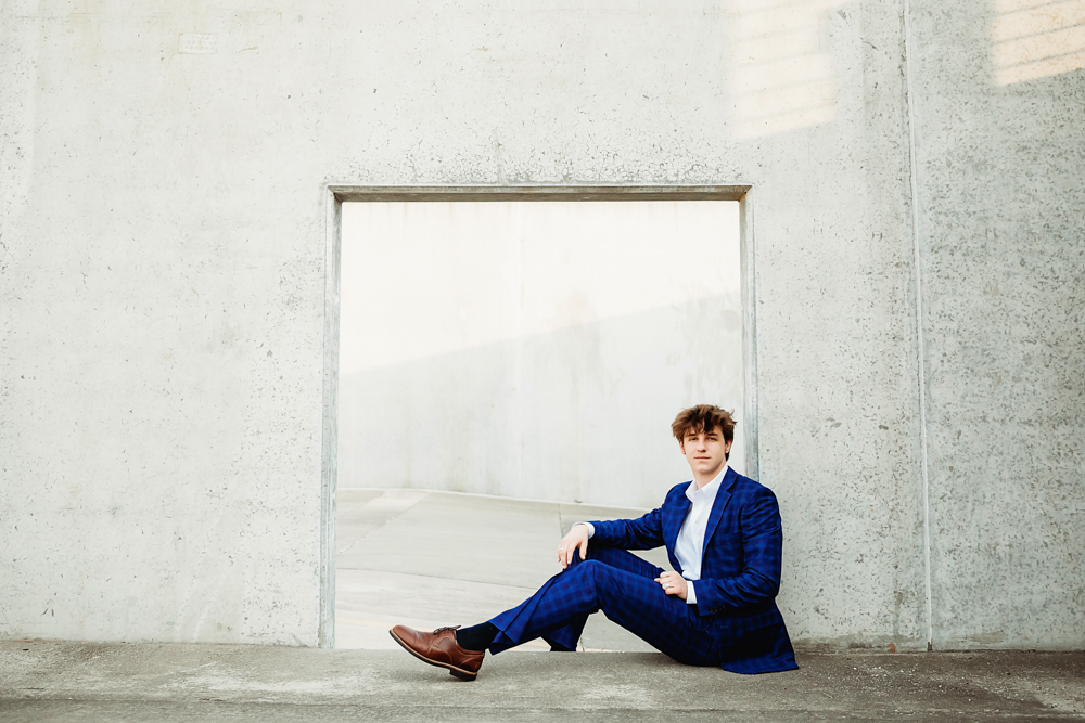 Jack seated in a modern concrete window opening wearing a sharp blue plaid suit and brown shoes in downtown Florence with Reflection Images by Tracy Rowell