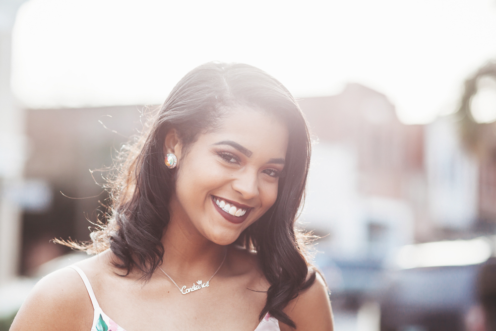Connie smiling brightly in golden hour light, wearing her floral dress and name necklace with soft curls at Reflection Images by Tracy Rowell in Florence SC