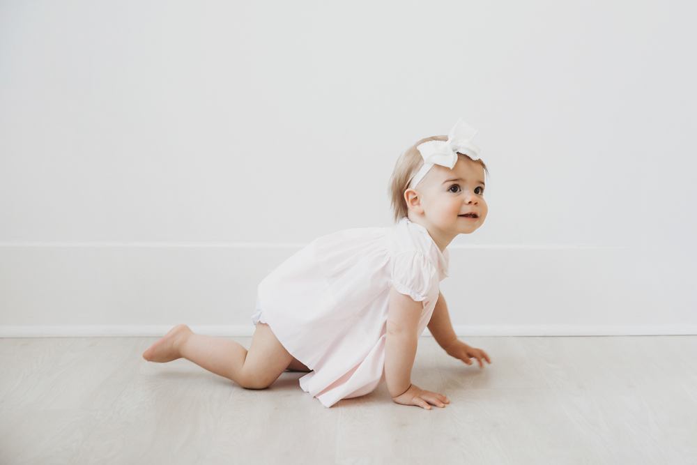 Grace crawling on light wood floors in a soft pink dress with a large white bow at Reflection Images by Tracy Rowell in Florence SC