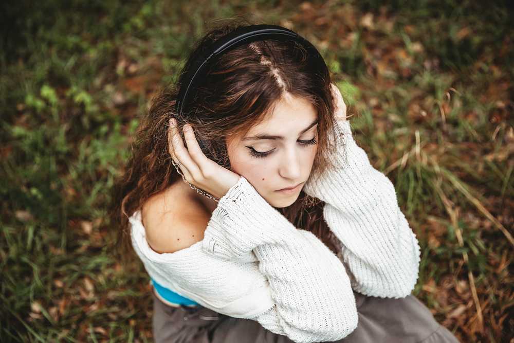 Eve with eyes closed wearing headphones, cream sweater slipping off shoulder, sitting in grass at Reflection Images by Tracy Rowell in Florence SC