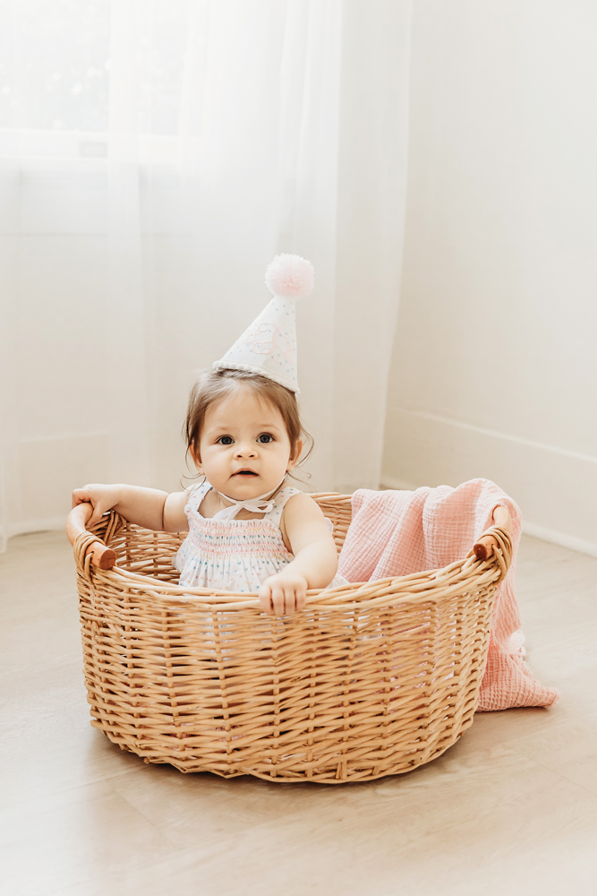 One-year-old girl sitting in a wicker basket with pink blanket, wearing birthday hat.