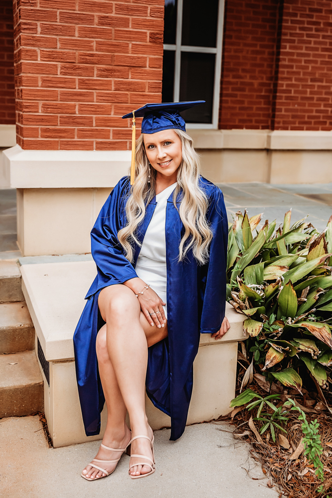 Kristina sitting gracefully on stone steps in her navy graduation gown with a brick wall and greenery in the background at Reflection Images by Tracy Rowell in Florence SC