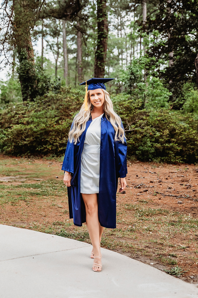 Full-length photo of Kristina smiling in a navy graduation gown on a path surrounded by trees at Reflection Images by Tracy Rowell in Florence SC
