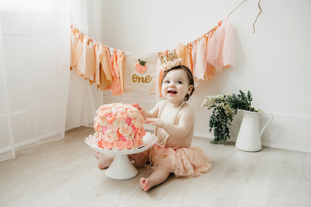 Aubrey in a peach tutu and gold crown sitting in front of a rosette cake and peach ONE banner at Reflection Images by Tracy Rowell in Florence SC