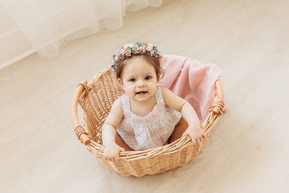 Birthday girl Livi Kate sits in a wicker basket with a floral crown and smiles during her studio photo session.