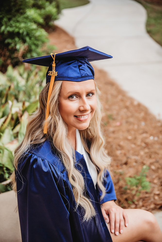 Close-up of Kristina smiling while seated in her cap and gown, with pine straw and a curving sidewalk behind at Reflection Images by Tracy Rowell in Florence SC