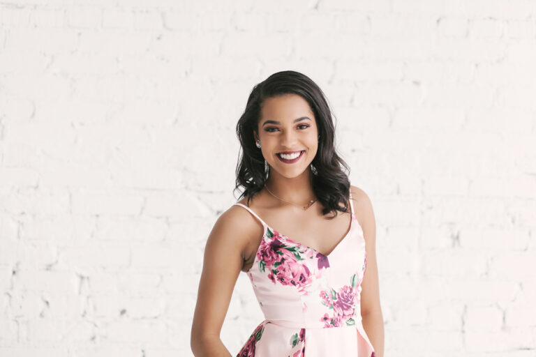 Indoor portrait of Connie with a soft smile against a white brick wall, wearing a pink floral dress and drop earrings at Reflection Images by Tracy Rowell in Florence SC