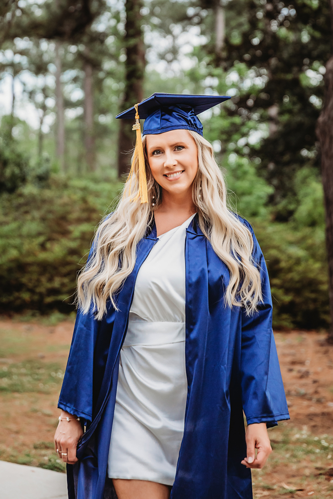 Mid-length shot of Kristina smiling in her gown and white dress, walking outdoors among pine trees at Reflection Images by Tracy Rowell in Florence SC