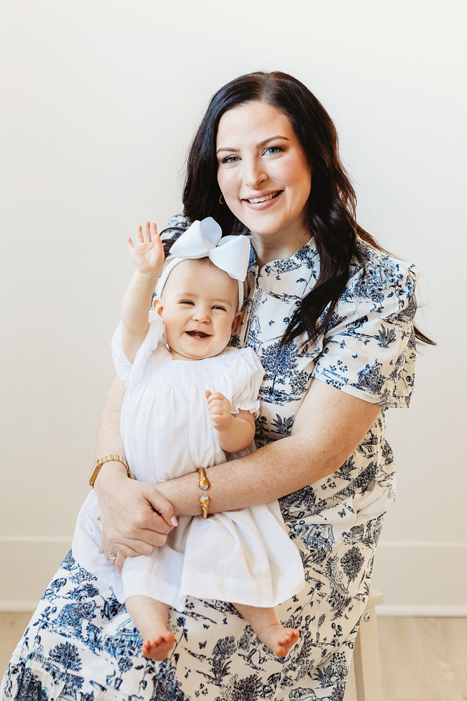 Elsie smiling with raised hand while sitting on mom’s lap, white dress and bow, soft backdrop at Reflection Images by Tracy Rowell in Florence SC