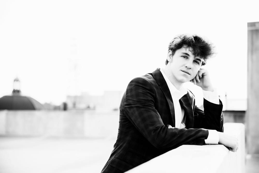 Black and white rooftop portrait of Jack resting on a ledge in his blue checkered suit with city skyline in the distance, Florence SC senior session