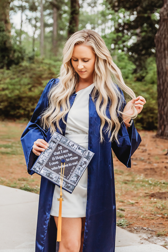 Kristina looking down at her decorated graduation cap with soft curls and navy gown in a pine grove at Reflection Images by Tracy Rowell in Florence SC