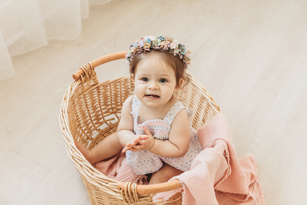 Livi Kate sits in a wicker basket with a pink blanket, wearing a pastel floral crown and looking curious.
