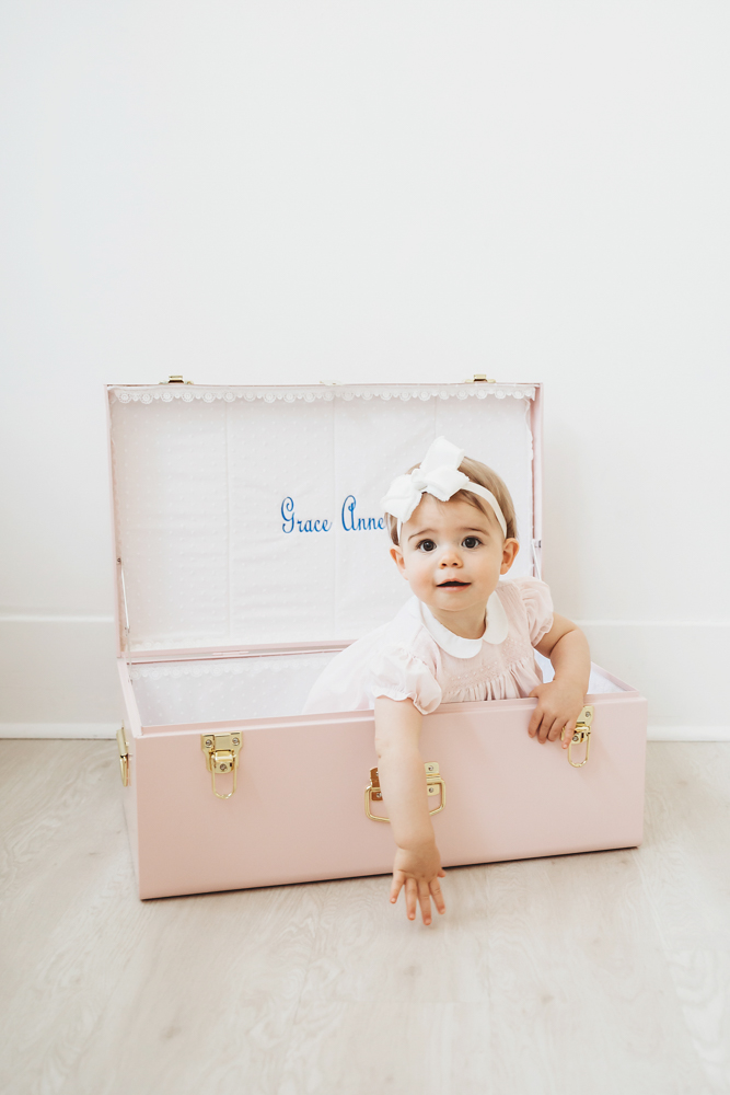 Grace peeking out from inside a personalized pink trunk that reads 'Grace Anne' in blue script at Reflection Images by Tracy Rowell in Florence SC