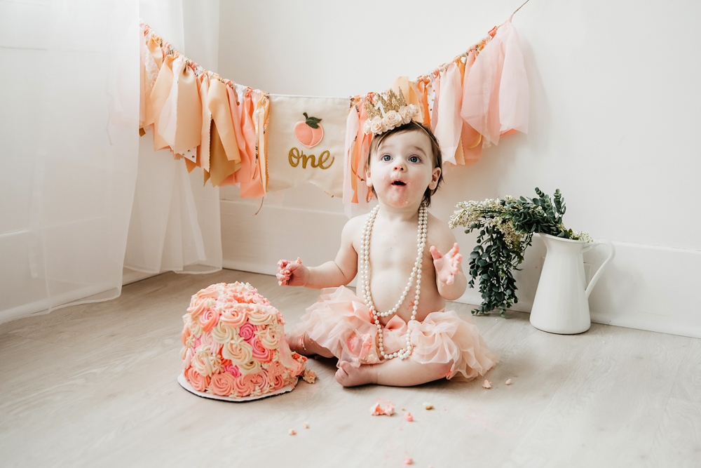 Aubrey with frosting on her hands sitting in front of a half-eaten pink cake and peach banner at Reflection Images by Tracy Rowell in Florence SC