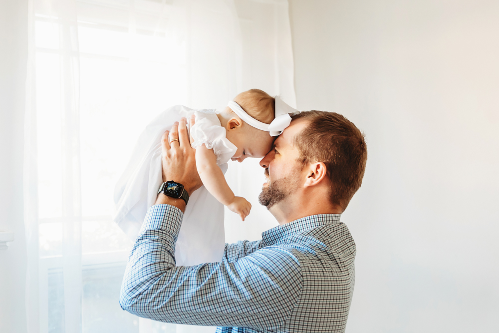 Sweet forehead-to-forehead moment with dad lifting Elsie in white dress and bow by the window at Reflection Images by Tracy Rowell in Florence SC