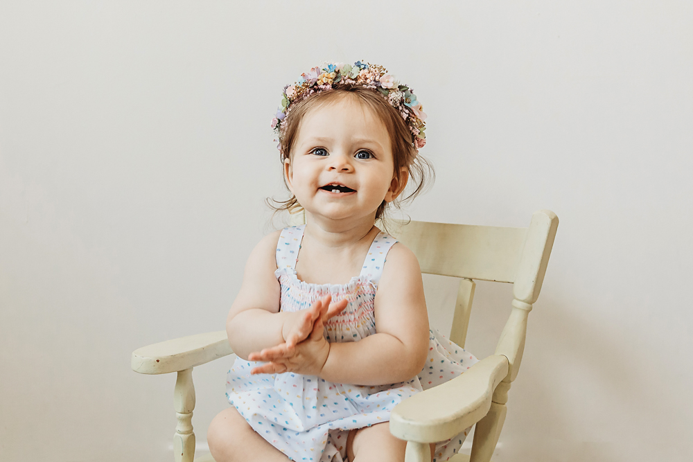 Livi Kate claps happily while sitting in a white wooden chair during her first birthday studio session.