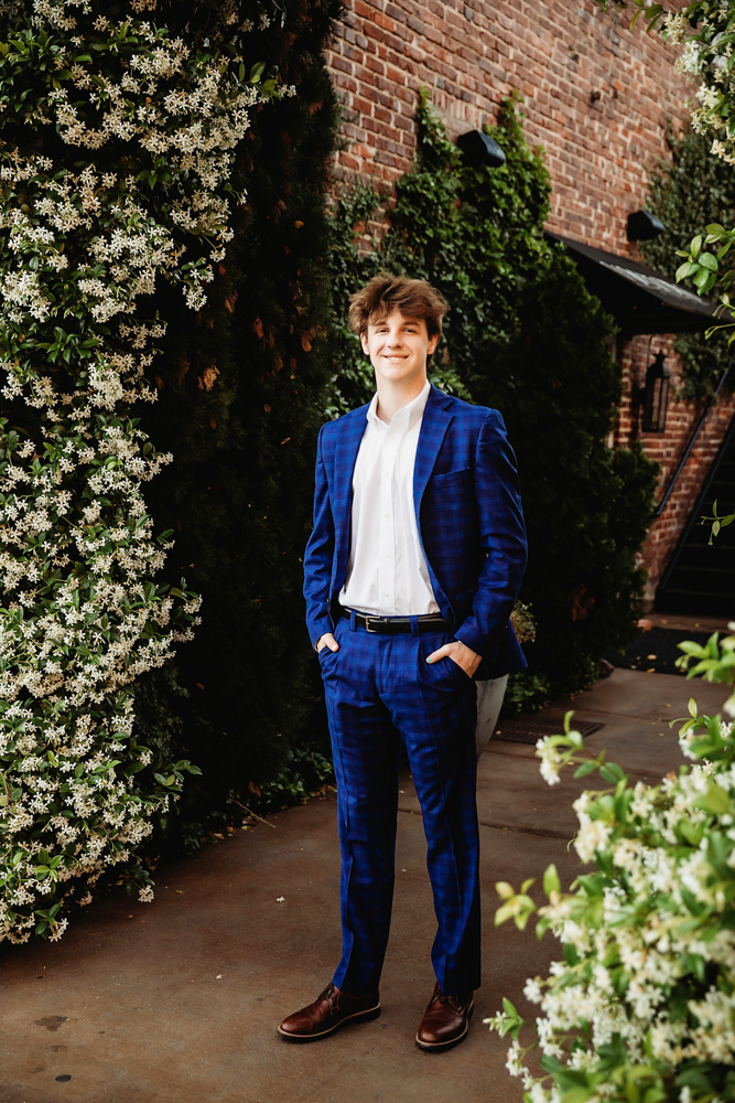 Full-body portrait of Jack standing between blooming white jasmine flowers and brick walls, wearing a blue suit and brown shoes in Florence SC