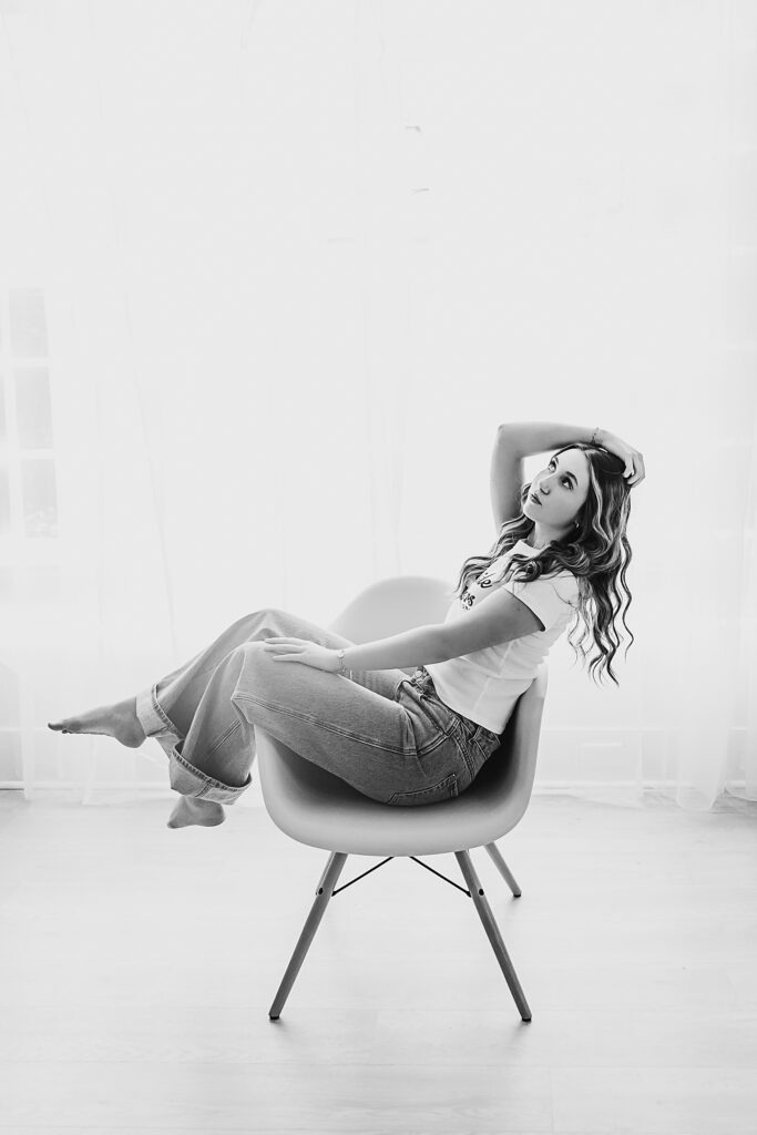 Black and white image of Addy lounging in a modern chair, arching back with arm in her hair, light flooding in from sheer curtains at Reflection Images by Tracy Rowell in Florence SC