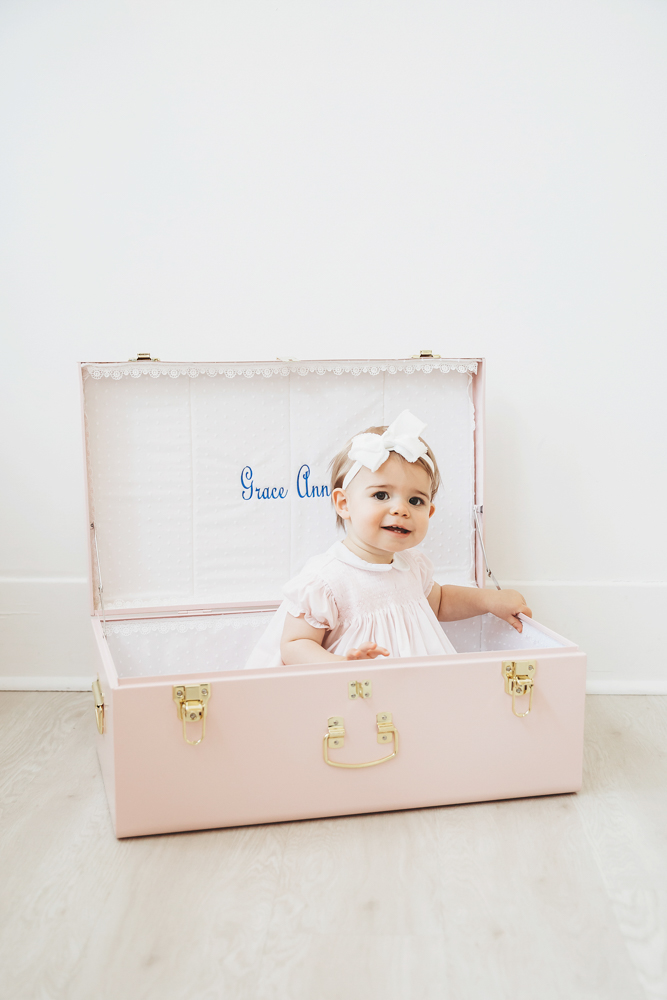 Grace sitting inside her pink name-embroidered trunk and smiling sweetly in a white dress at Reflection Images by Tracy Rowell in Florence SC