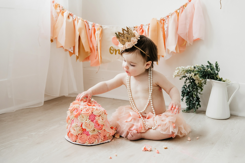 Aubrey reaching toward the rosette cake in a tutu and crown, pearls draped around her, during her cake smash at Reflection Images by Tracy Rowell in Florence SC
