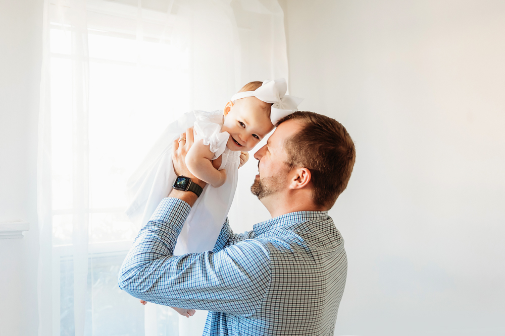 Dad lifting Elsie with big smile, white bow and dress, bright window light at Reflection Images by Tracy Rowell in Florence SC