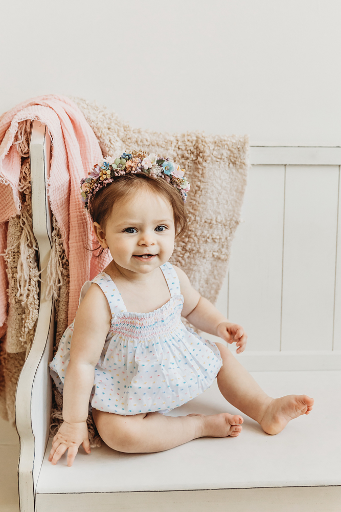 Birthday girl Livi Kate poses on a white bench with soft blankets and a flower crown during her studio session.
