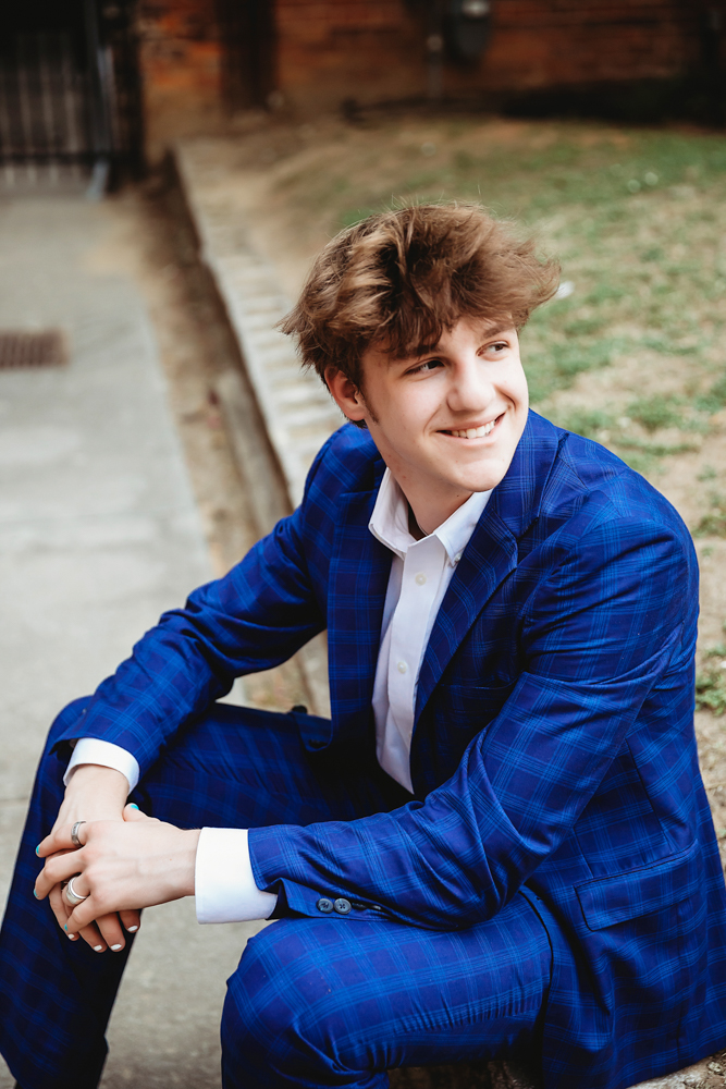 Jack sitting on a curb with a soft smile, wearing a tailored blue plaid suit and white shirt, captured near brick and grass textures in downtown Florence SC