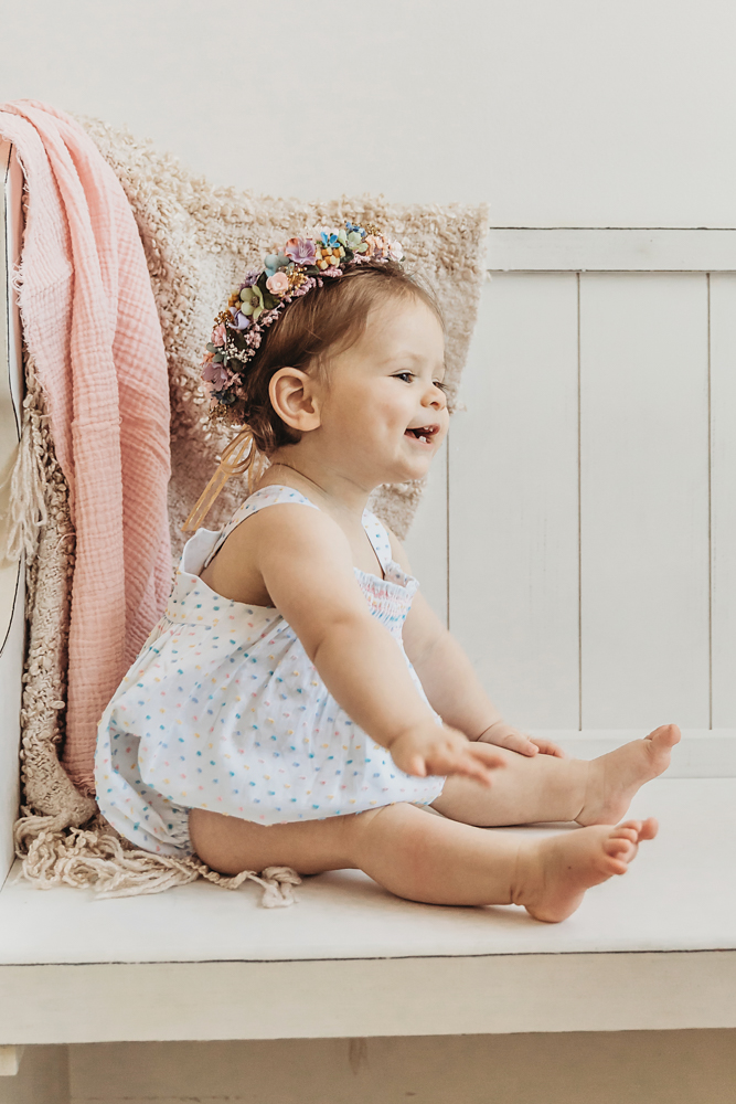 Livi Kate sits on a cozy bench, smiling and wearing a multicolor floral crown and pastel dress.