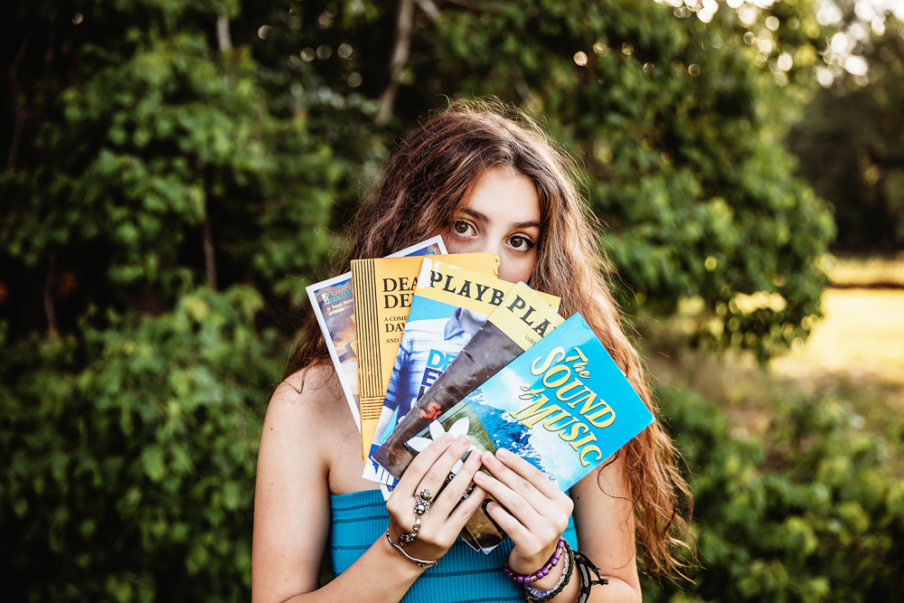 Eve holding colorful theater playbills covering her mouth, turquoise bandeau visible, greenery in background at Reflection Images by Tracy Rowell in Florence SC