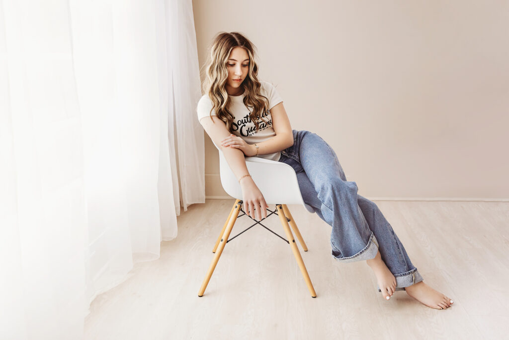 Teen portrait of Addy in a relaxed pose on a white chair, looking down thoughtfully with wavy hair and soft shadows at Reflection Images by Tracy Rowell in Florence SC