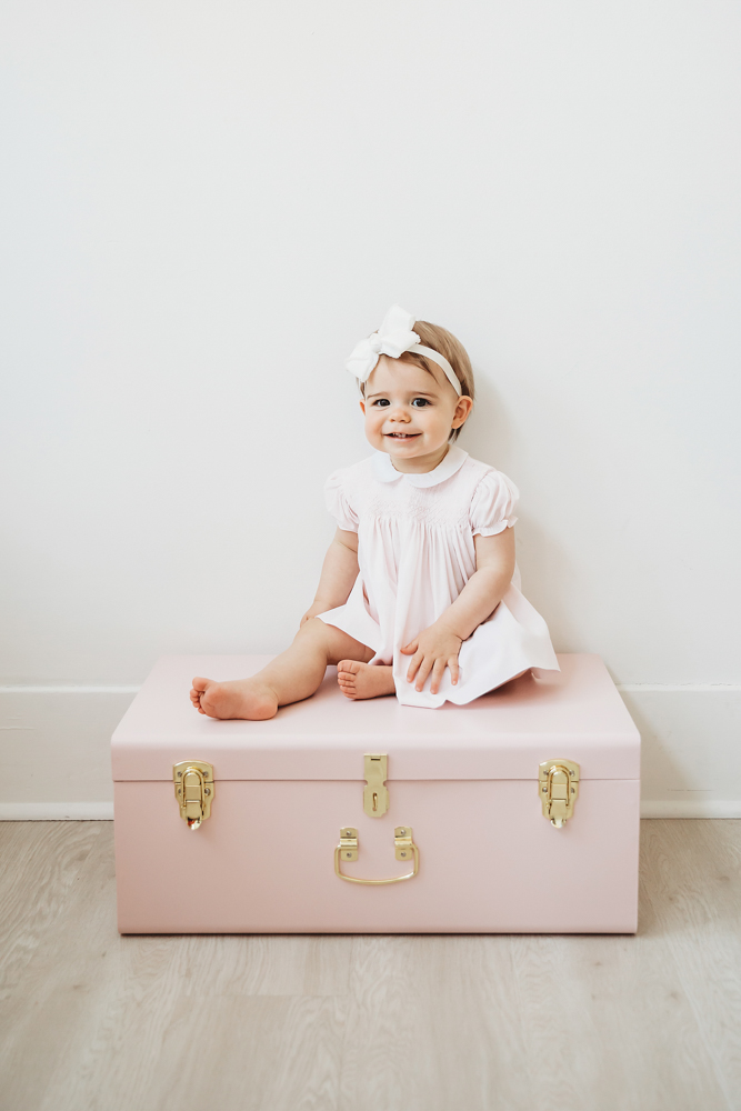 Grace perched barefoot on top of a closed pink trunk against a white wall, wearing a pale pink dress and white bow at Reflection Images by Tracy Rowell in Florence SC